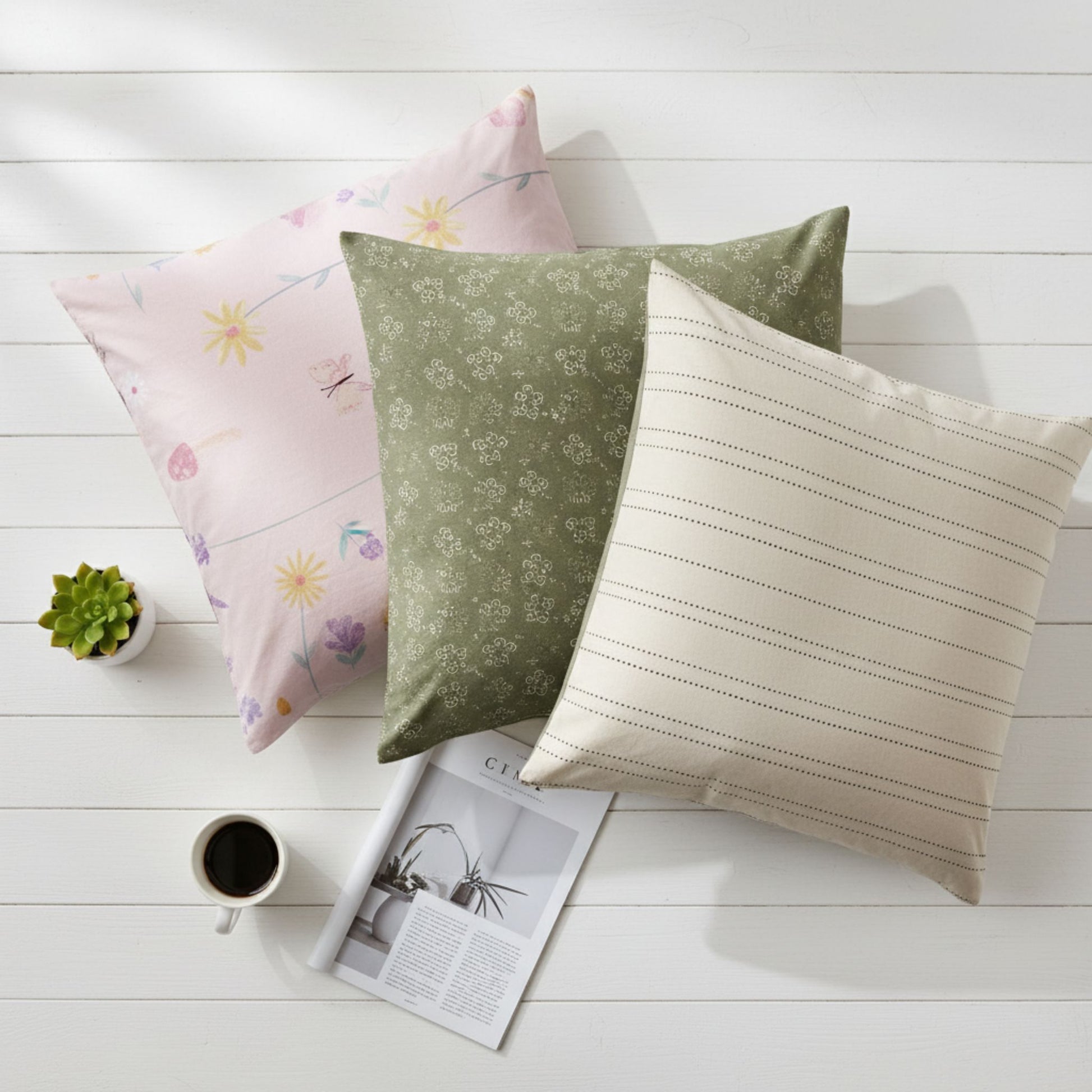 Three decorative pillow cases on a white wooden surface with a coffee cup and plant, featuring ZM Textiles Belgium printed pillowcase set.
